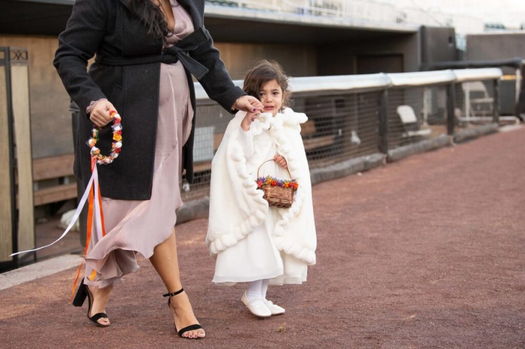 Heather-Scott-Wedding-Low-Res-73 1 - TOMMY GATZ ENTERTAINMENT A young girl wearing a white coat and holding a basket walks hand in hand with an adult in a dress and coat along a dirt path.