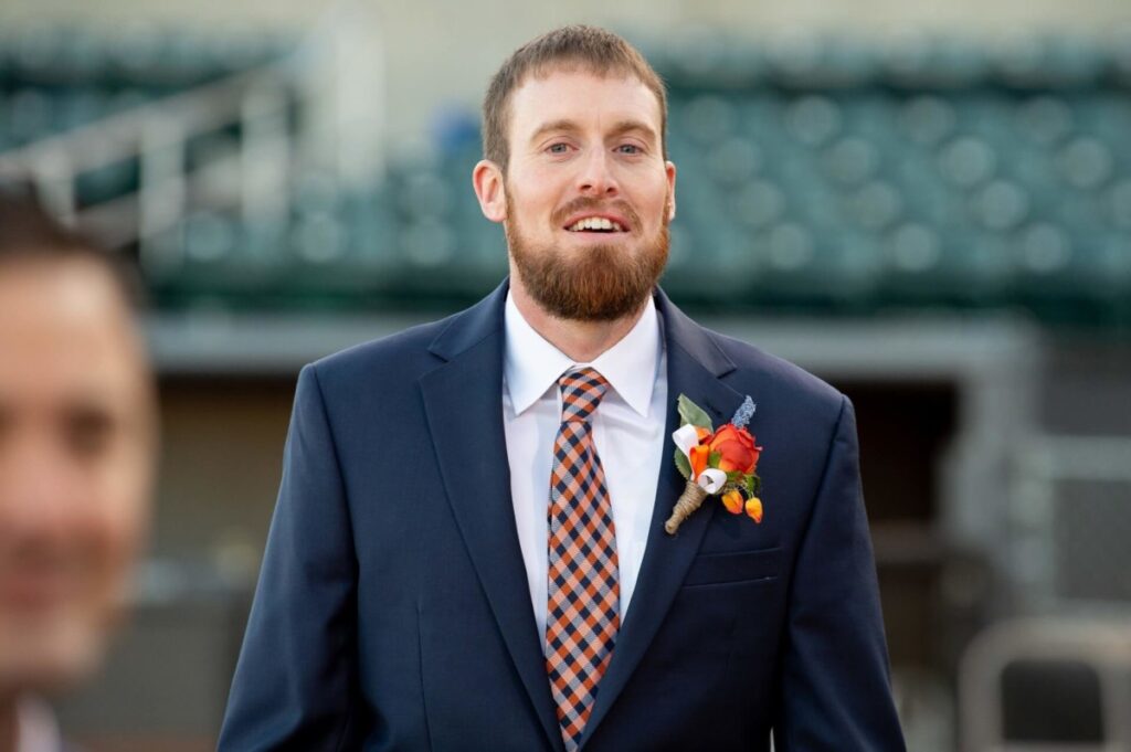 Heather-Scott-Wedding-Low-Res-74 1 - TOMMY GATZ ENTERTAINMENT A bearded man in a suit with an orange and blue tie stands outside, wearing a boutonniere with orange flowers.