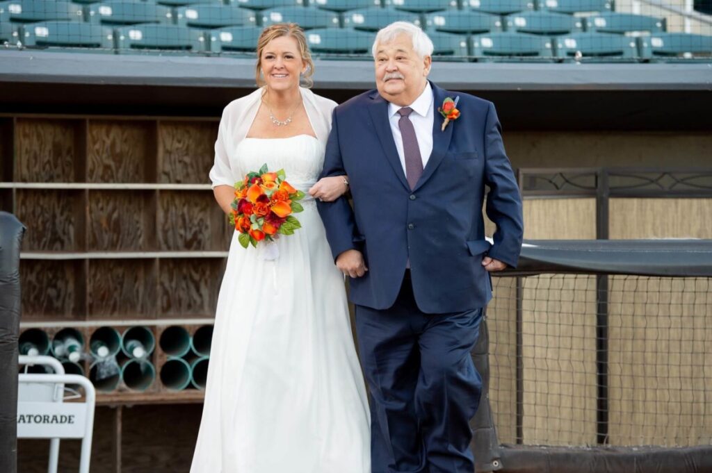 Heather-Scott-Wedding-Low-Res-75 1 - TOMMY GATZ ENTERTAINMENT A bride in a white dress holds a bouquet of orange flowers while standing beside an older man in a blue suit. They are outdoors, near a dugout at a stadium with empty bleachers.