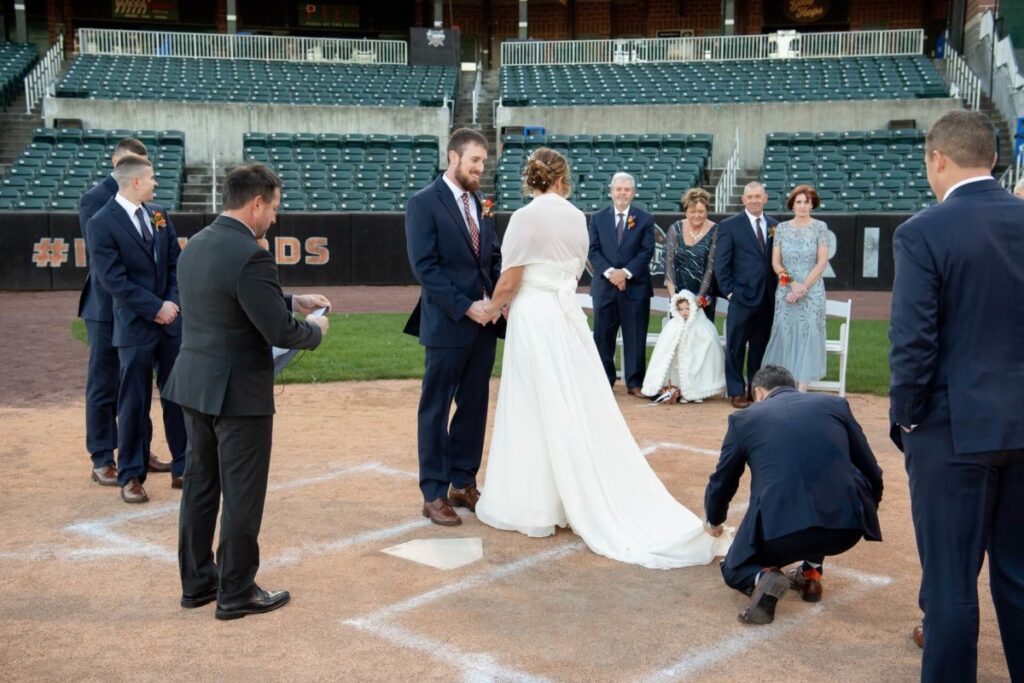 Heather-Scott-Wedding-Low-Res-76 1 - TOMMY GATZ ENTERTAINMENT A couple exchanges vows in a baseball stadium, surrounded by their wedding party and family. One person kneels to adjust the bride's dress.