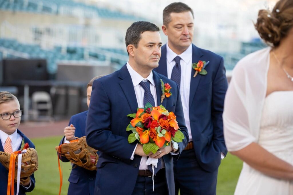 Heather-Scott-Wedding-Low-Res-78 1 - TOMMY GATZ ENTERTAINMENT Two men in suits hold colorful bouquets at an outdoor event, with boys holding baseball gloves nearby.