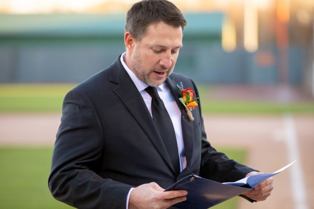 Heather-Scott-Wedding-Low-Res-79 1 - TOMMY GATZ ENTERTAINMENT A man in a suit reads from a folder outdoors, wearing a boutonniere on his lapel.