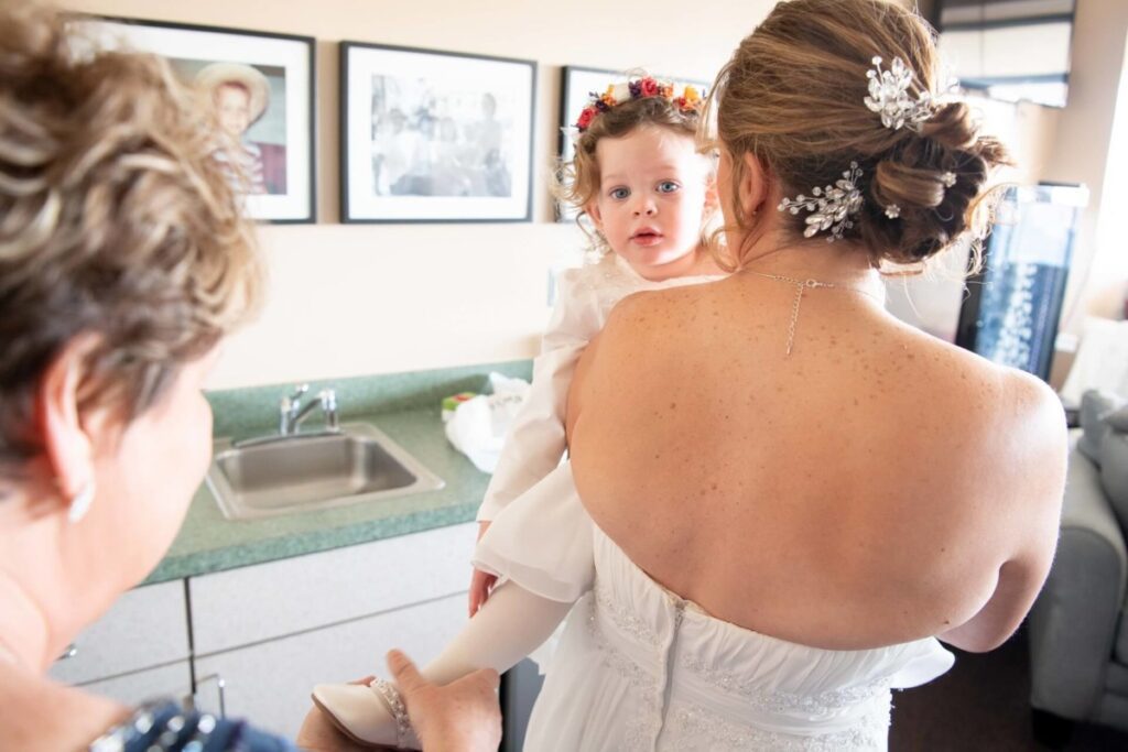 Heather-Scott-Wedding-Low-Res-8 1 - TOMMY GATZ ENTERTAINMENT A woman in a white dress holds a young child with curly hair and a flower crown, in a room with framed photos on the wall.