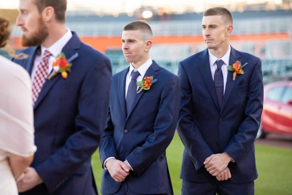 Heather-Scott-Wedding-Low-Res-80 1 - TOMMY GATZ ENTERTAINMENT Three men in blue suits stand outdoors at an event. Two have boutonnières and are looking ahead.