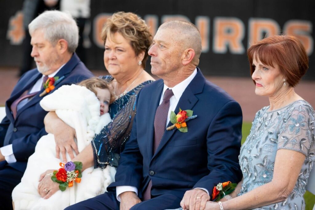 Heather-Scott-Wedding-Low-Res-81 1 - TOMMY GATZ ENTERTAINMENT Four adults seated, dressed formally, with corsages. One holds a bundled child. They're outdoors with a blurred "BIRDS" sign in the background.