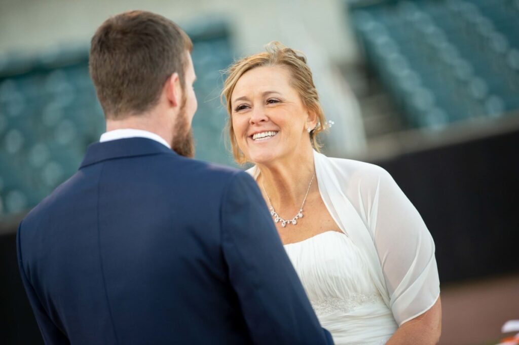 Heather-Scott-Wedding-Low-Res-82 1 - TOMMY GATZ ENTERTAINMENT A couple, in formal attire, stands outdoors. The woman smiles at the man, who has his back to the camera. They are in a setting with green and white seats in the background.