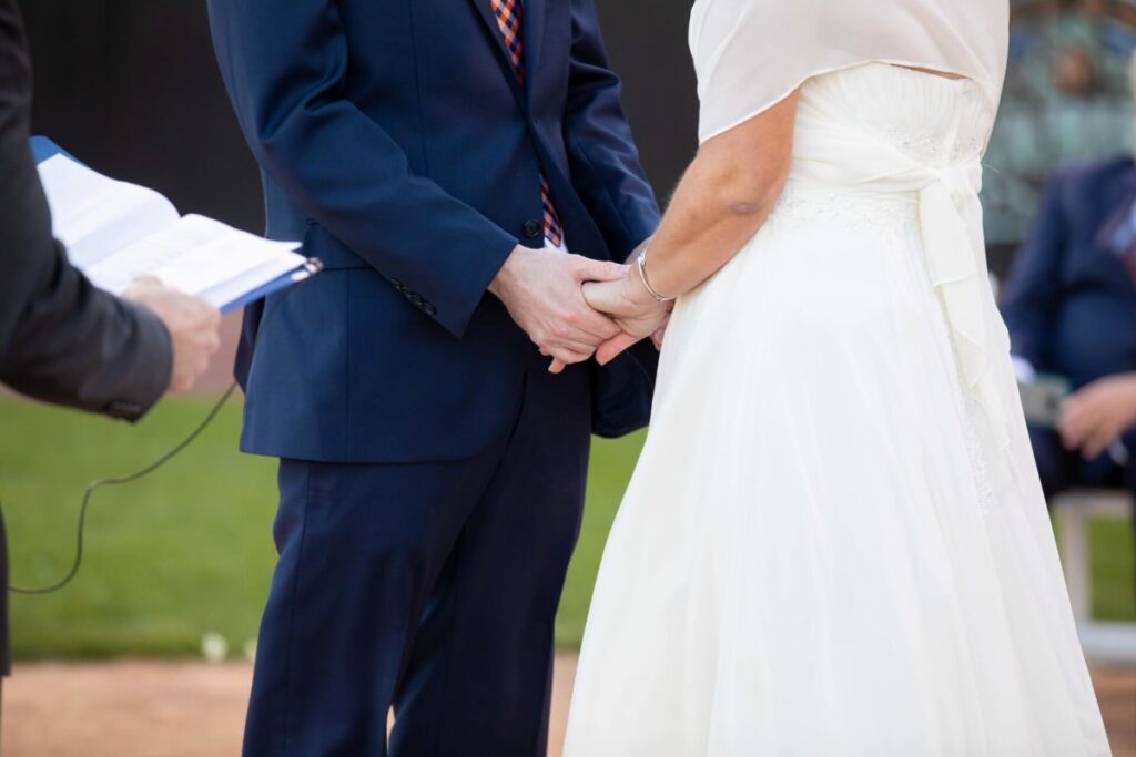 Heather-Scott-Wedding-Low-Res-83 1 - TOMMY GATZ ENTERTAINMENT A couple in wedding attire holds hands during a ceremony, with a person holding papers standing nearby.