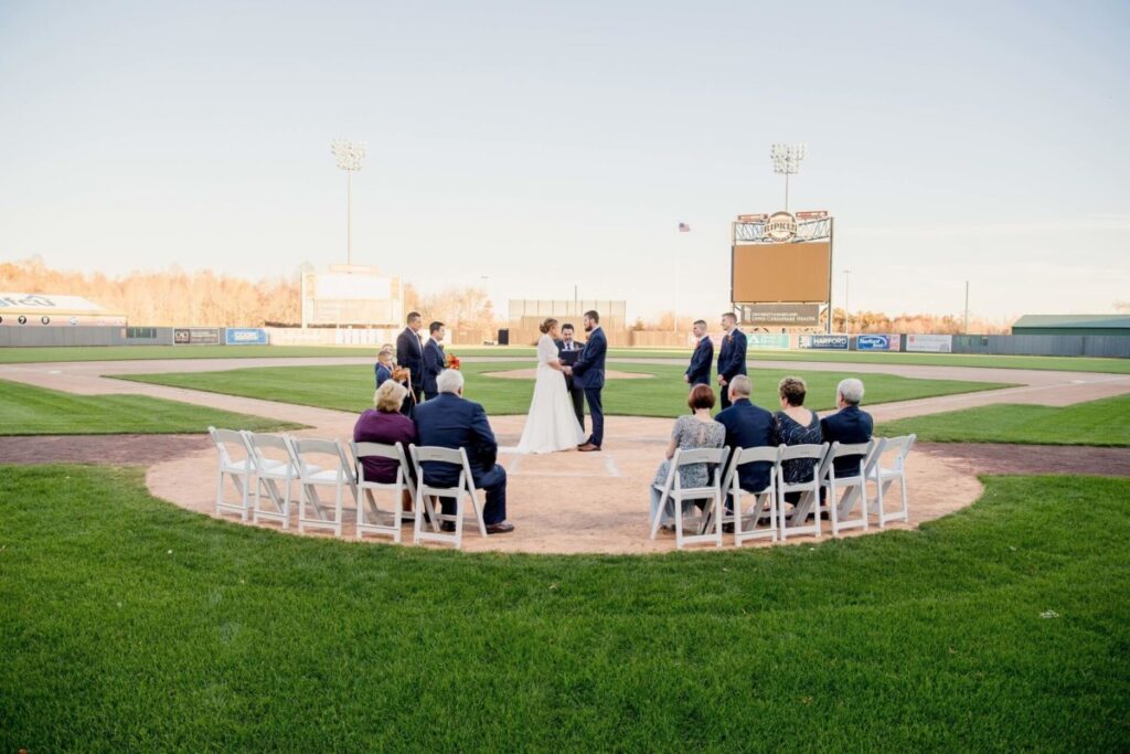 Heather-Scott-Wedding-Low-Res-85 1 - TOMMY GATZ ENTERTAINMENT A wedding ceremony is taking place on a baseball field, with the couple standing on the pitcher's mound, surrounded by a small gathering of seated guests.