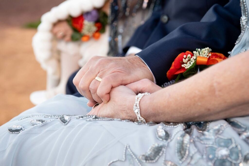 Heather-Scott-Wedding-Low-Res-86 1 - TOMMY GATZ ENTERTAINMENT A couple holding hands, dressed in formal attire with detailed embroidery, wearing jewelry. A bouquet with red flowers is visible in the background.