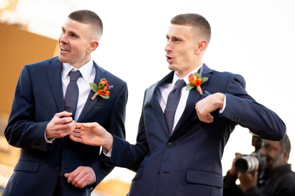 Heather-Scott-Wedding-Low-Res-87 1 - TOMMY GATZ ENTERTAINMENT Two men in suits, wearing boutonnières, stand outdoors gesturing animatedly. A photographer is visible in the background.