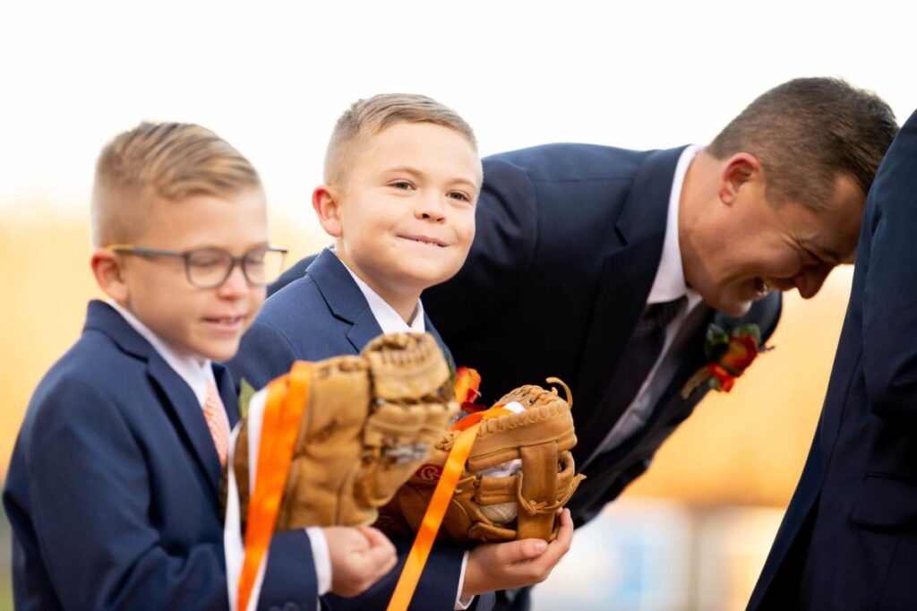 Heather-Scott-Wedding-Low-Res-88 1 - TOMMY GATZ ENTERTAINMENT Two boys in suits hold baseball gloves with ribbons, standing next to a suited man who is laughing.