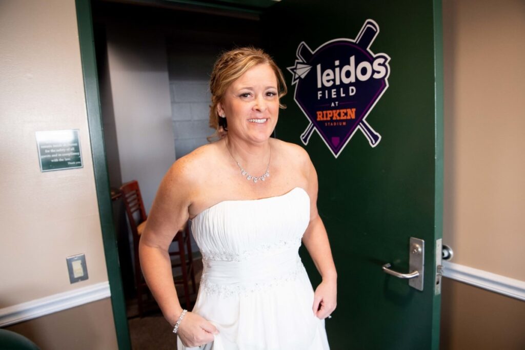 Heather-Scott-Wedding-Low-Res-9 1 - TOMMY GATZ ENTERTAINMENT A woman in a white strapless dress stands in a room with a green door, which has a "Leidos Field at Ripken Stadium" sign.