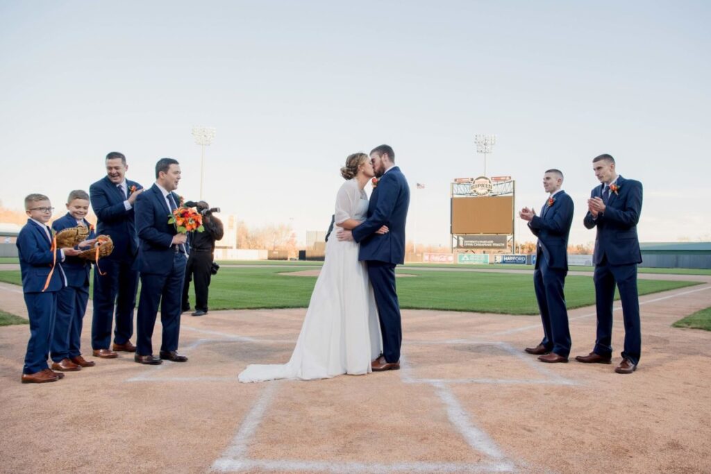 Heather-Scott-Wedding-Low-Res-90 1 - TOMMY GATZ ENTERTAINMENT A couple is getting married on a baseball field. They are kissing at home plate surrounded by the bridal party, all dressed in formal attire.