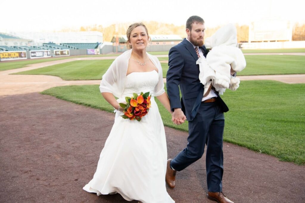 Heather-Scott-Wedding-Low-Res-92 1 - TOMMY GATZ ENTERTAINMENT Bride and groom walking on a baseball field, holding hands. The bride holds a bouquet, and the groom carries a baby swaddled in a blanket.