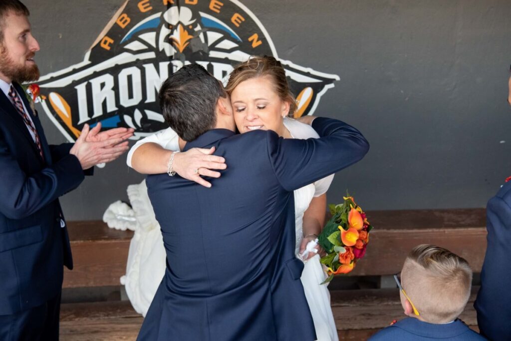 Heather-Scott-Wedding-Low-Res-93 1 - TOMMY GATZ ENTERTAINMENT Bride hugging a man in a suit, holding an orange bouquet. Two men and a child in suits are nearby. They stand in front of an Aberdeen IronBirds logo.