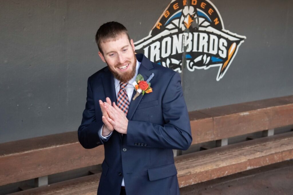 Heather-Scott-Wedding-Low-Res-94 1 - TOMMY GATZ ENTERTAINMENT Man in a suit with a boutonniere stands smiling in front of an "Aberdeen IronBirds" logo in a dugout.