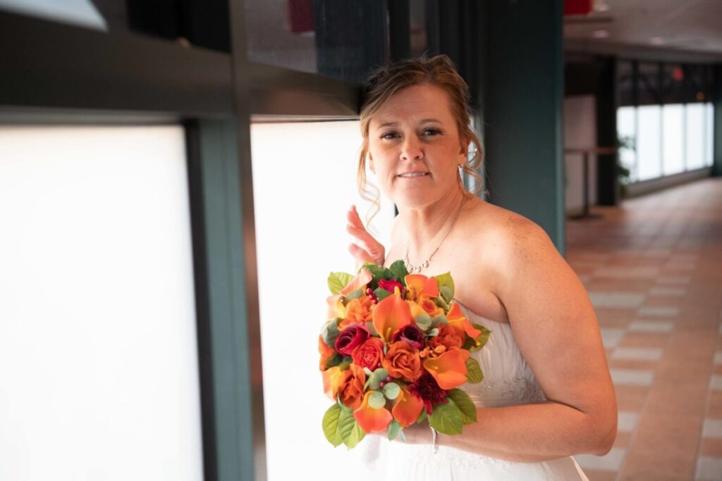 Heather-Scott-Wedding-Low-Res-96 1 - TOMMY GATZ ENTERTAINMENT A bride holding a bouquet of orange and red flowers stands indoors near a window, wearing a white dress.