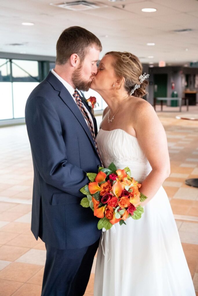 Heather-Scott-Wedding-Low-Res-98 1 - TOMMY GATZ ENTERTAINMENT A bride and groom share a kiss indoors. The bride holds a bouquet of orange and red flowers, and the groom is in a navy suit.