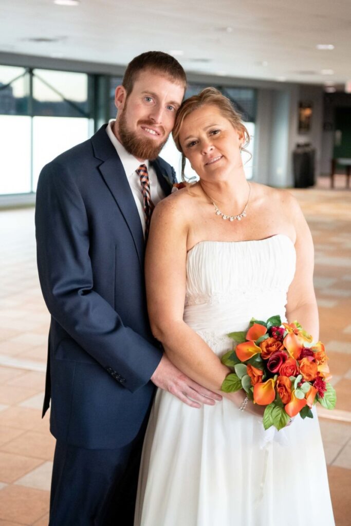 Heather-Scott-Wedding-Low-Res-99 1 - TOMMY GATZ ENTERTAINMENT A couple dressed in wedding attire stands indoors. The bride holds a bouquet of orange and red flowers.