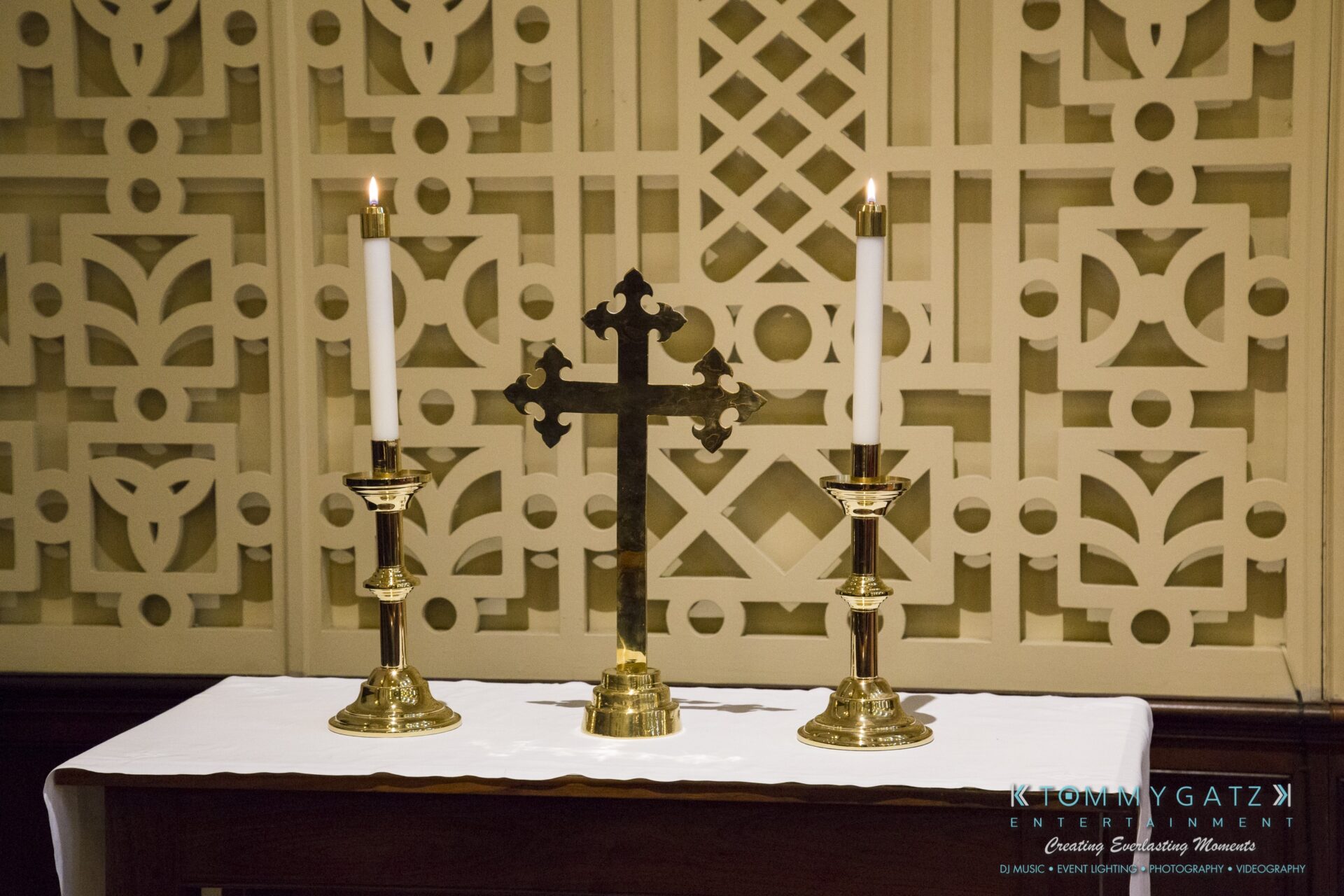 Jamie-and-Mark-Low-Res-Watermark-100 - TOMMY GATZ ENTERTAINMENT Two lit candles and a decorative cross on a white altar cloth in front of a patterned wall.