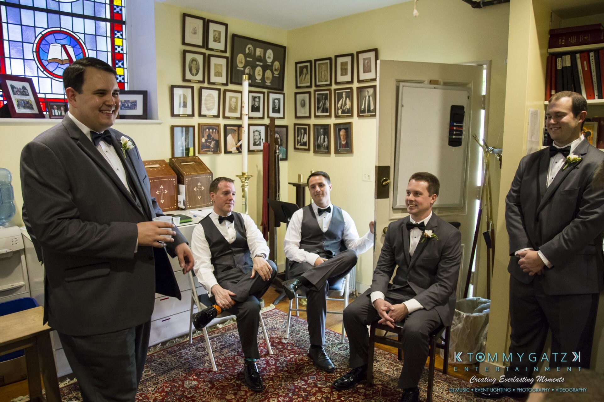 Jamie-and-Mark-Low-Res-Watermark-107 - TOMMY GATZ ENTERTAINMENT A group of five men in suits and tuxedos are gathered in a room with framed pictures on the wall, a water cooler, and a rug. Three are seated, and two are standing and smiling.