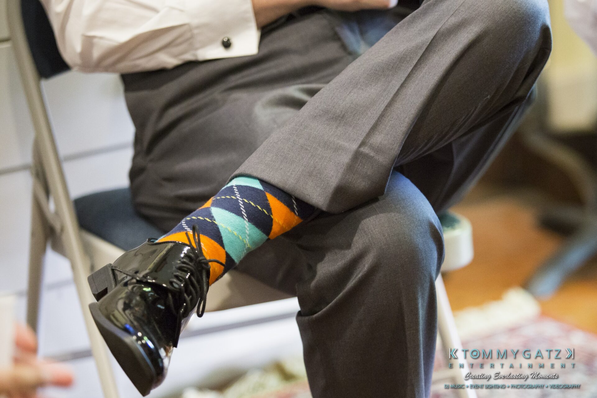 Jamie-and-Mark-Low-Res-Watermark-109 - TOMMY GATZ ENTERTAINMENT Person wearing gray pants and black shoes with colorful argyle socks, sitting on a folding chair.