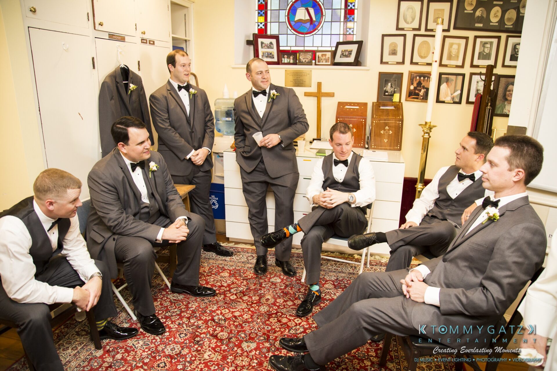 Jamie-and-Mark-Low-Res-Watermark-110 - TOMMY GATZ ENTERTAINMENT A group of seven men in formal attire sit and stand in a room with framed pictures and a stained glass window.