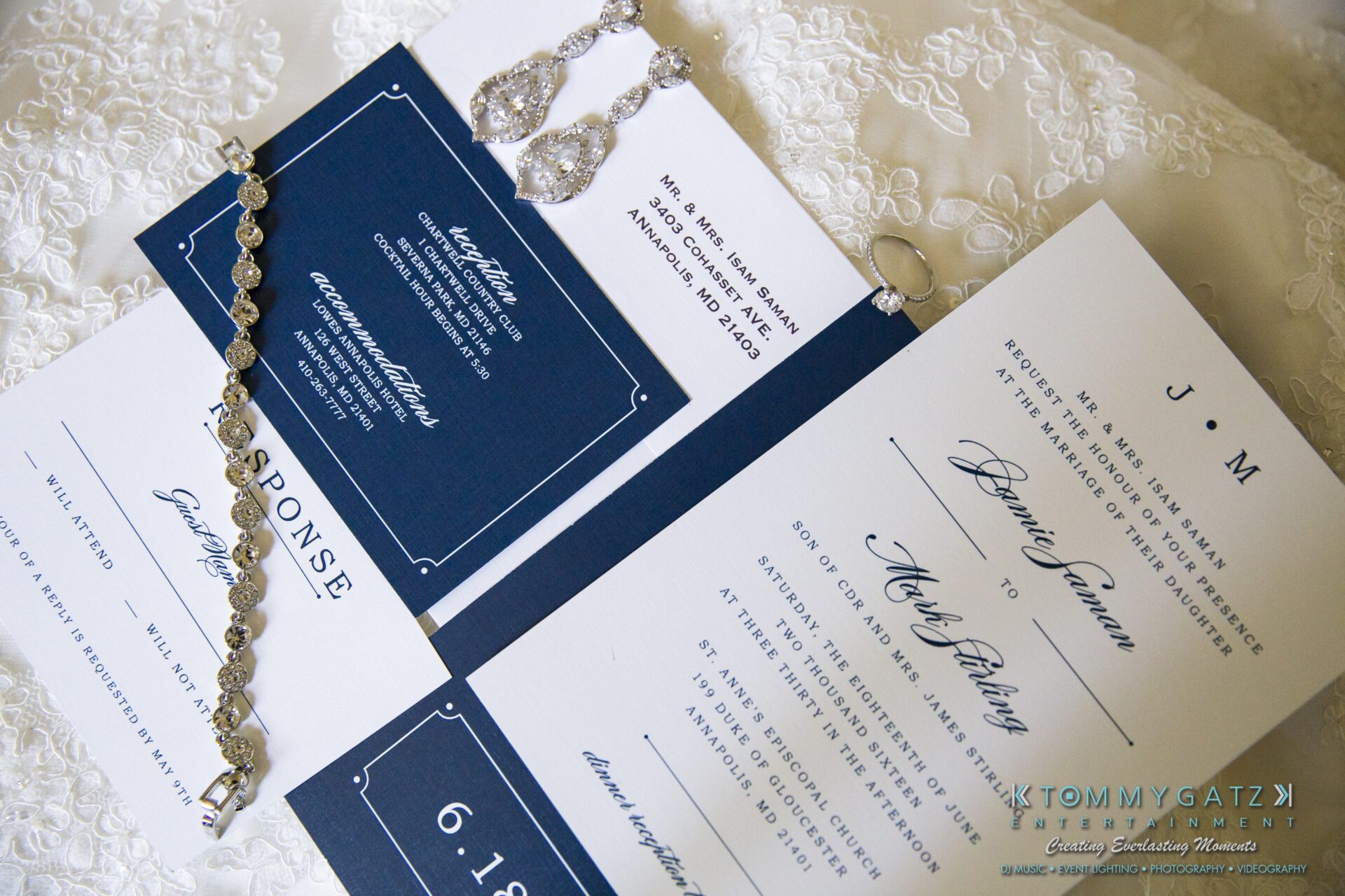 Jamie-and-Mark-Low-Res-Watermark-13 - TOMMY GATZ ENTERTAINMENT A navy and white wedding invitation suite displayed on lace fabric, adorned with a bracelet and earrings. The date on the invite is June 19.