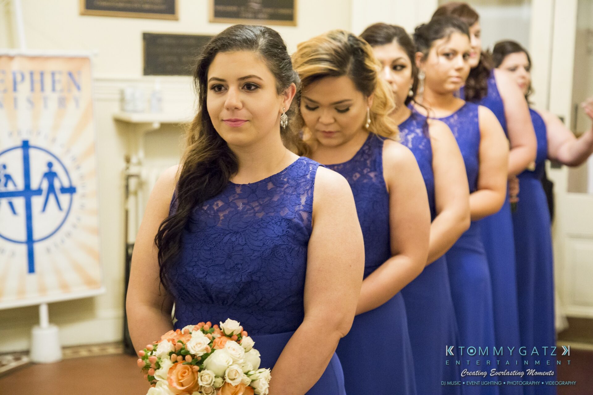Jamie-and-Mark-Low-Res-Watermark-132 - TOMMY GATZ ENTERTAINMENT A line of women in matching blue dresses stand inside a room. The woman in front holds a bouquet of flowers.