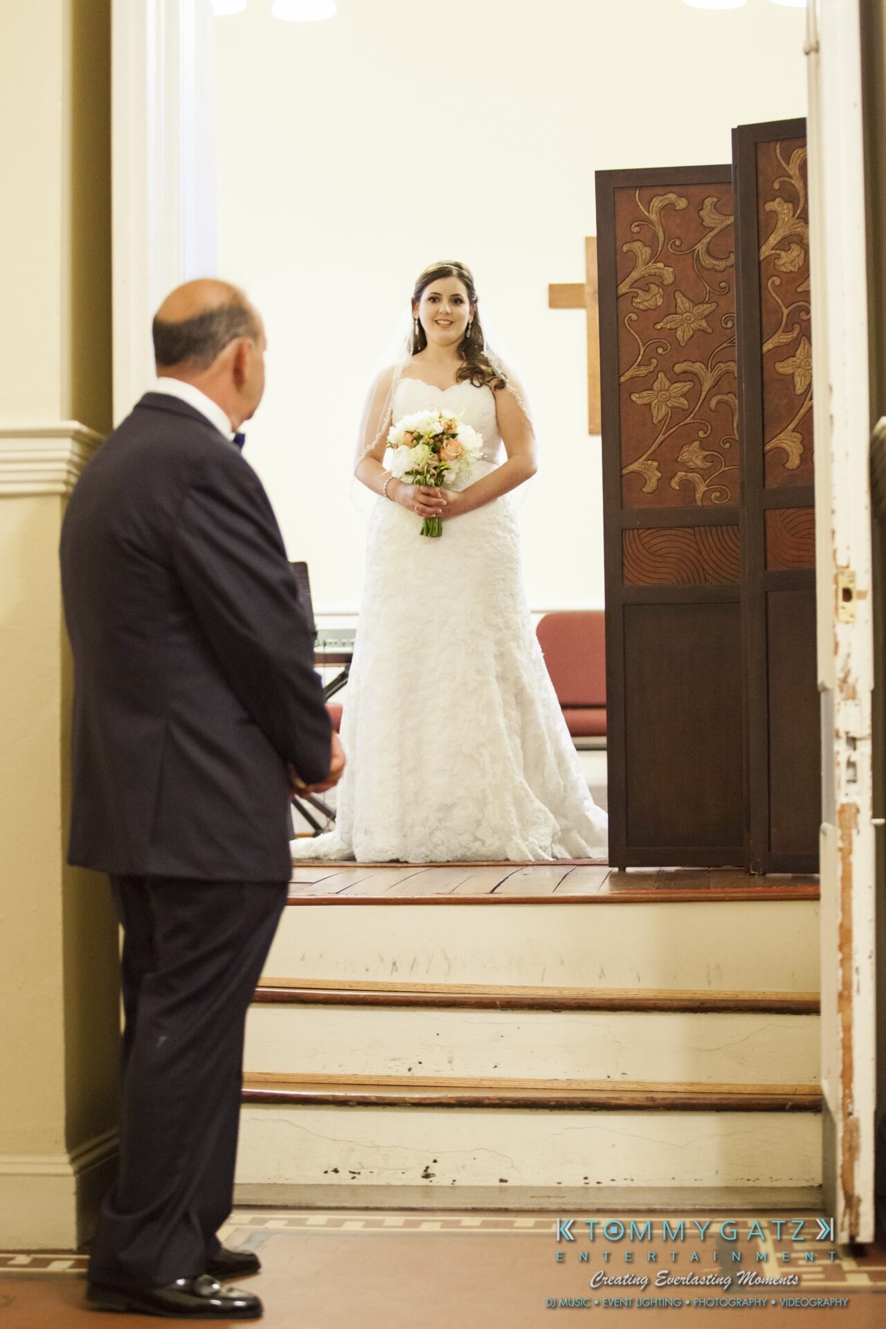 Jamie-and-Mark-Low-Res-Watermark-149 - TOMMY GATZ ENTERTAINMENT Bride in a white dress stands at the top of a short set of steps, holding a bouquet. A man in a suit stands at the bottom, facing her. A decorative panel is partially visible on the right.