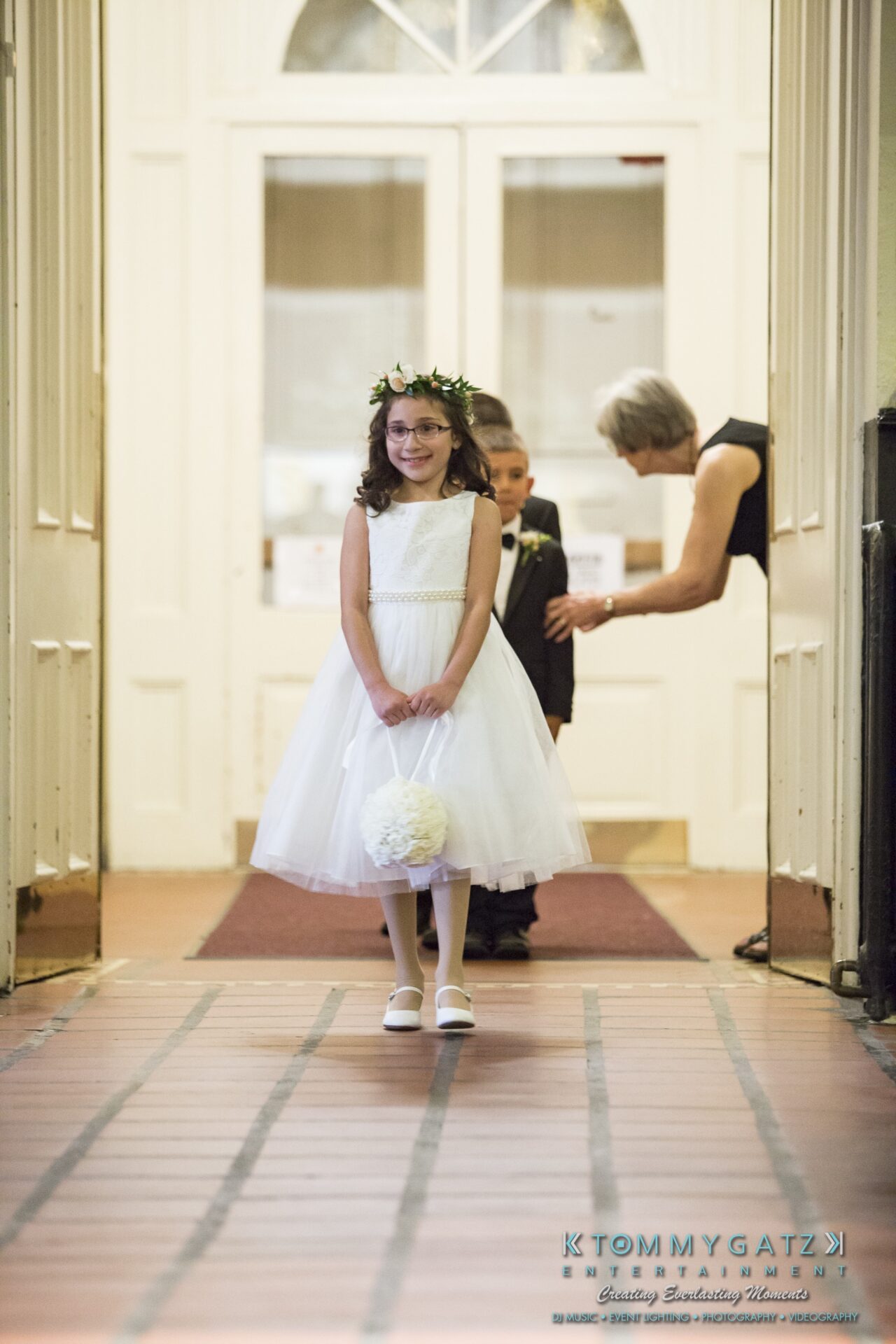Jamie-and-Mark-Low-Res-Watermark-164 - TOMMY GATZ ENTERTAINMENT A young flower girl in a white dress and floral crown walks down an aisle, holding a bouquet, with a young boy and an adult in the background.
