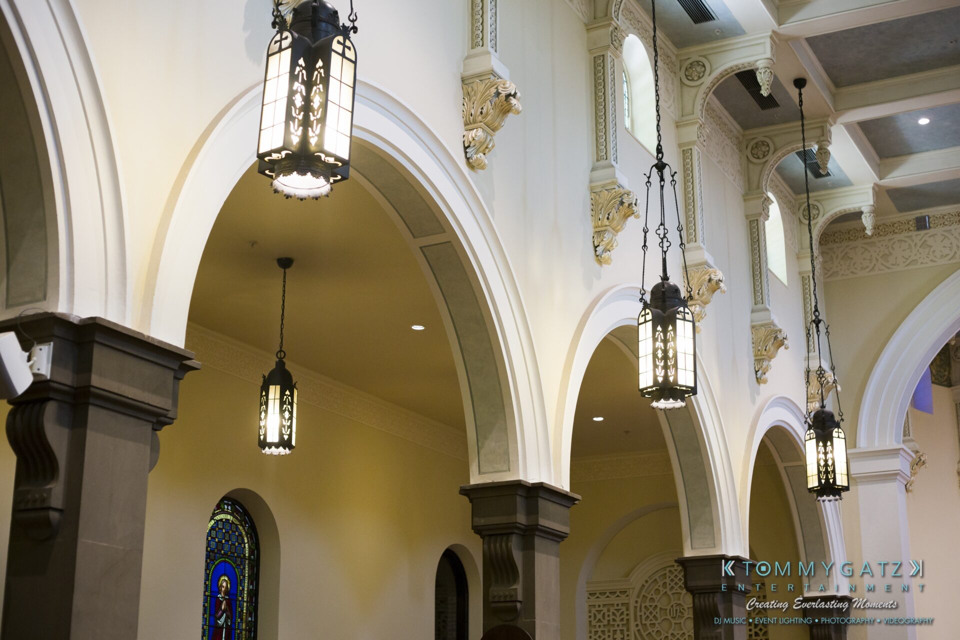 Jamie-and-Mark-Low-Res-Watermark-17 - TOMMY GATZ ENTERTAINMENT The interior of a building with ornate arched architecture and hanging lanterns. Stained glass windows are visible in the background.