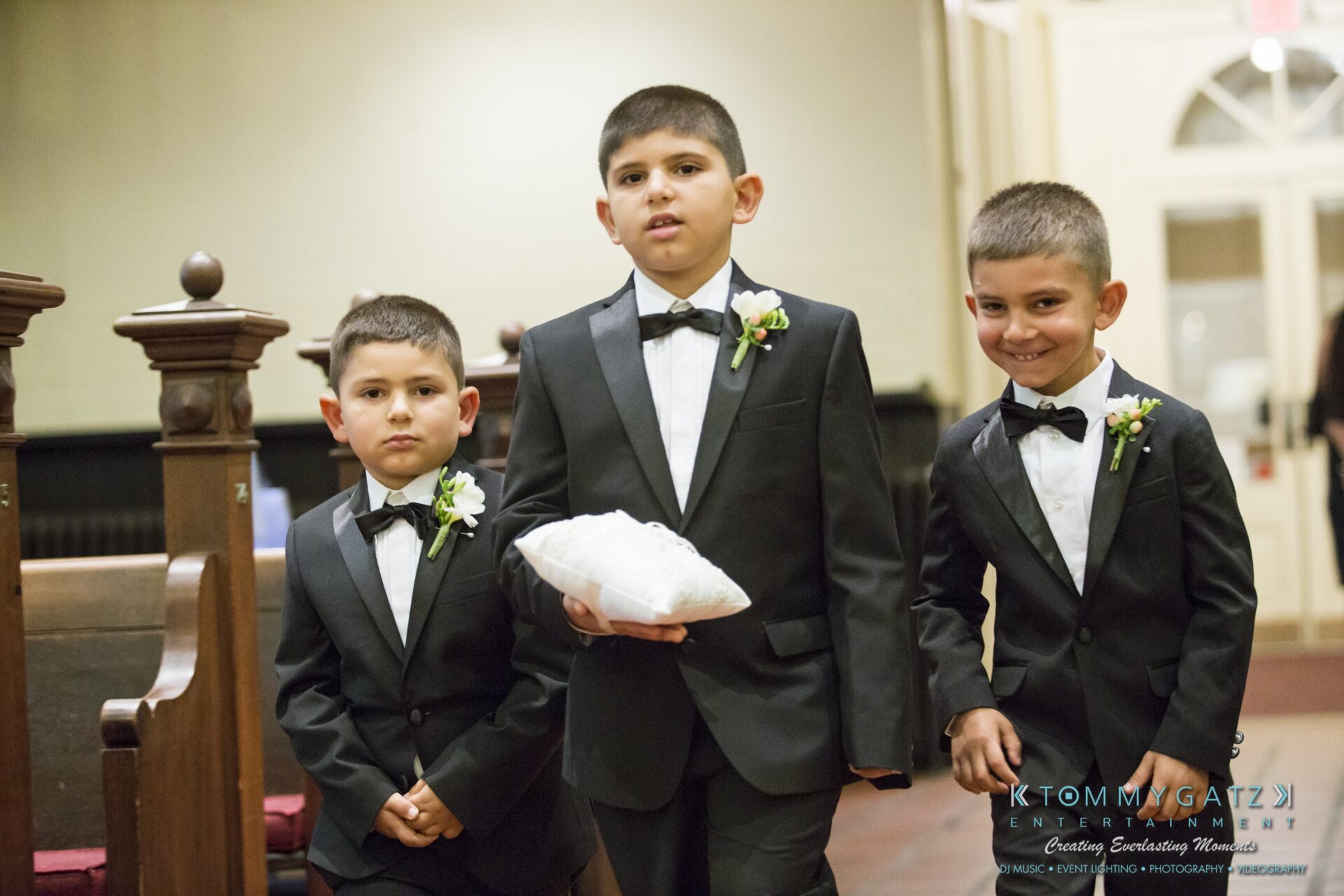 Jamie-and-Mark-Low-Res-Watermark-173 - TOMMY GATZ ENTERTAINMENT Three boys in black suits with bow ties and white boutonnieres, one holding a pillow, walk down the aisle in a church setting.