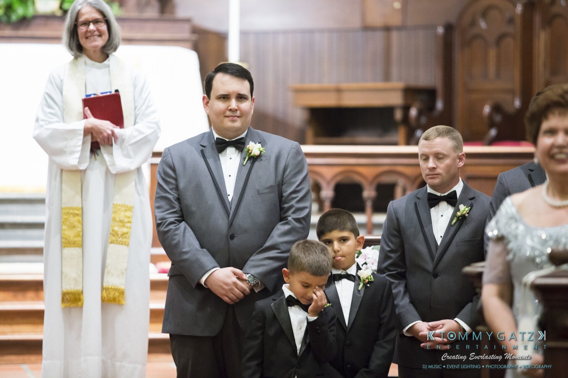 Jamie-and-Mark-Low-Res-Watermark-178 - TOMMY GATZ ENTERTAINMENT Wedding ceremony with a groom and two young boys in tuxedos. A smiling officiant stands nearby holding a book.
