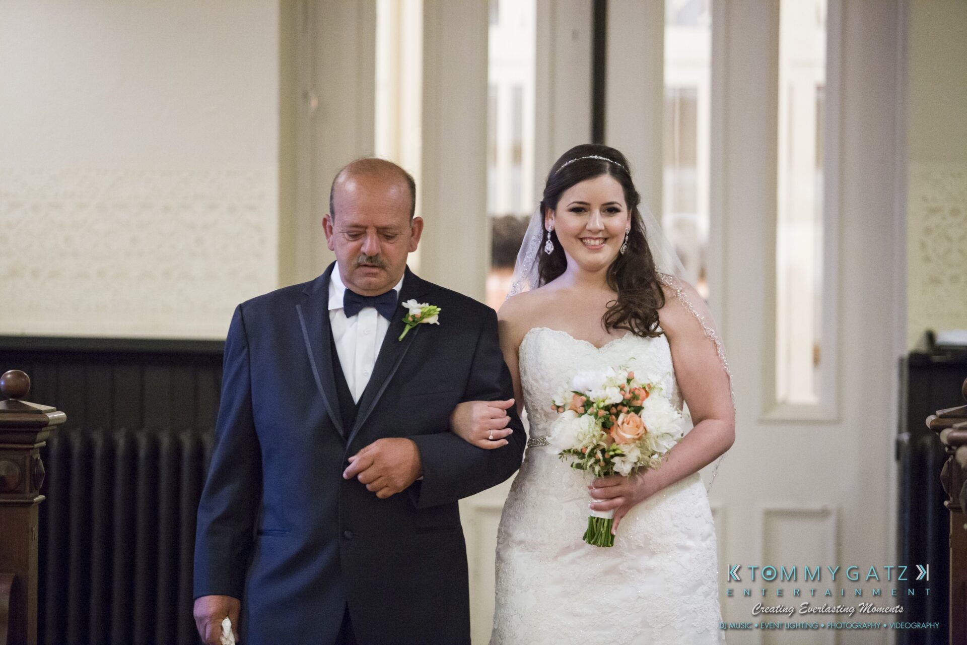 Jamie-and-Mark-Low-Res-Watermark-186 - TOMMY GATZ ENTERTAINMENT A bride in a white gown and veil walks down the aisle with a man in a tuxedo, holding a bouquet of flowers.