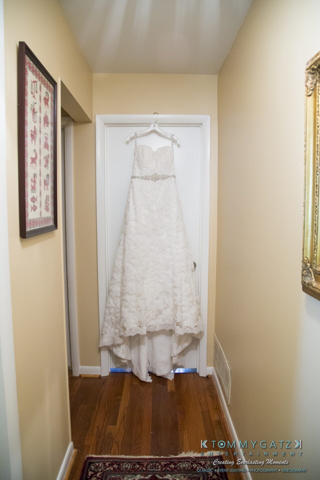Jamie-and-Mark-Low-Res-Watermark-20 - TOMMY GATZ ENTERTAINMENT A white lace wedding dress hangs on a door in a narrow, beige hallway with framed pictures on the walls.