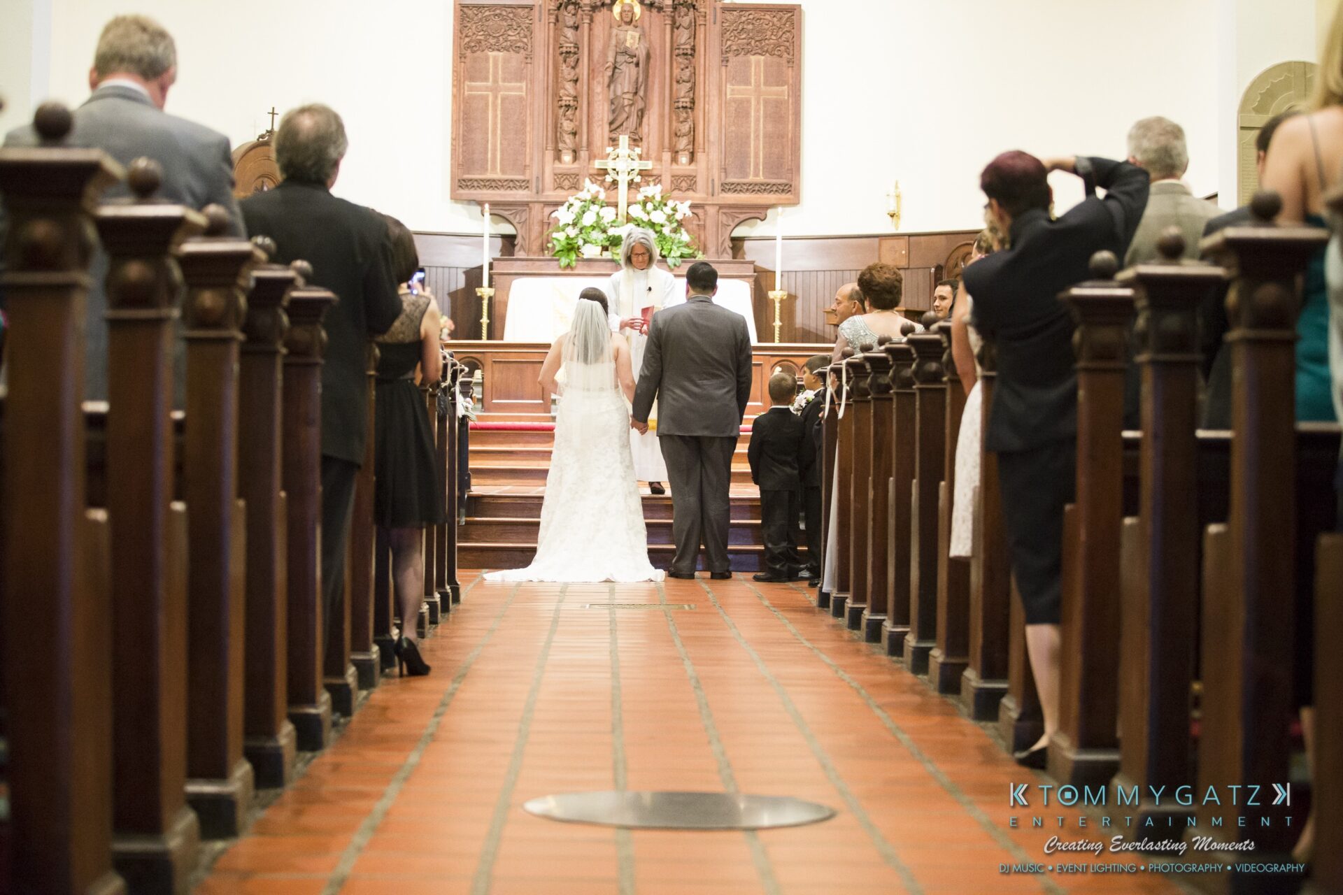 Jamie-and-Mark-Low-Res-Watermark-200 - TOMMY GATZ ENTERTAINMENT Wedding ceremony in a church with a couple standing at the altar, officiant in the background, and guests seated on both sides.