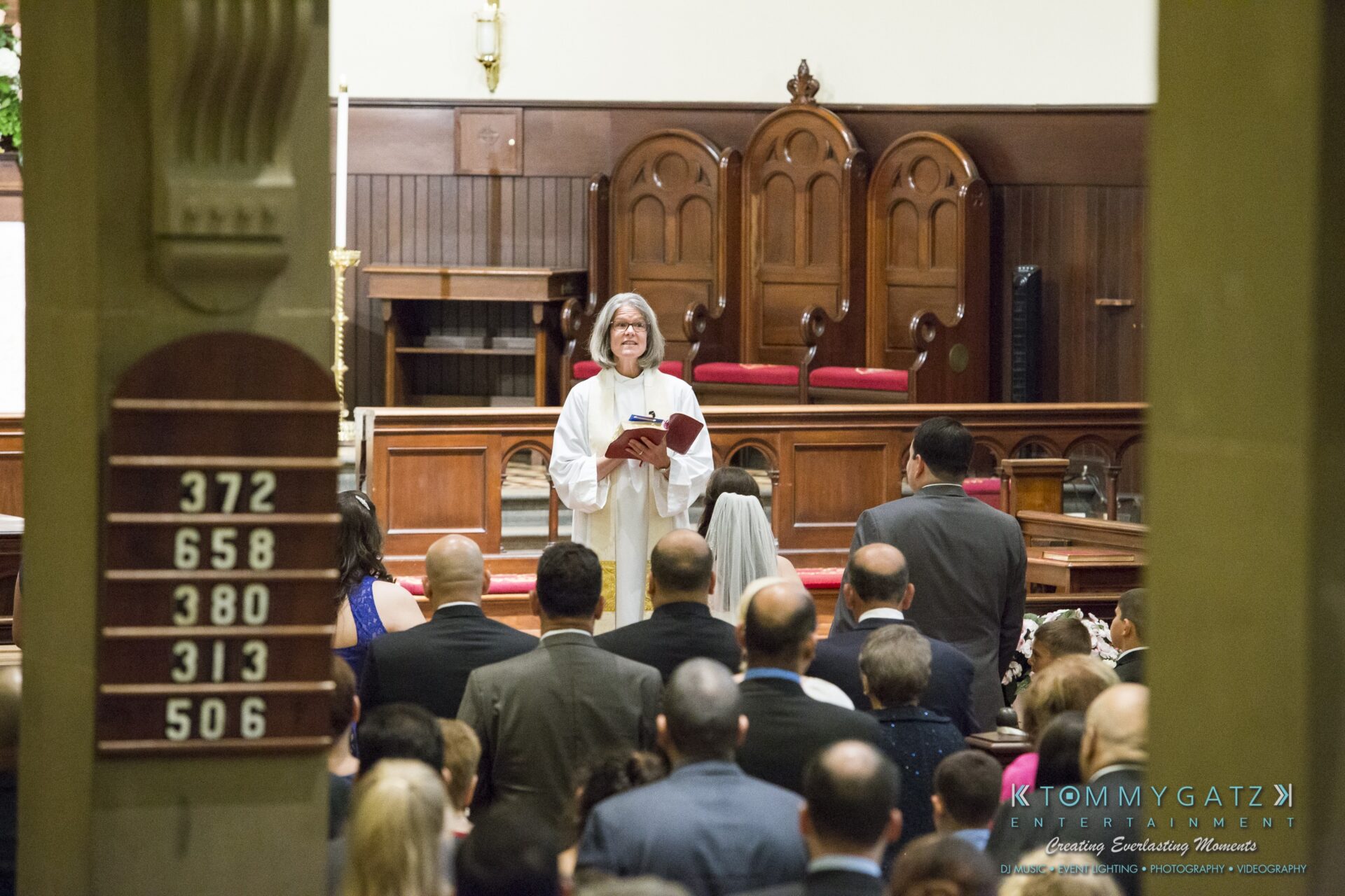 Jamie-and-Mark-Low-Res-Watermark-208 - TOMMY GATZ ENTERTAINMENT A priest stands at the altar holding a book, addressing a congregation in a church. People are seated and standing in the pews.