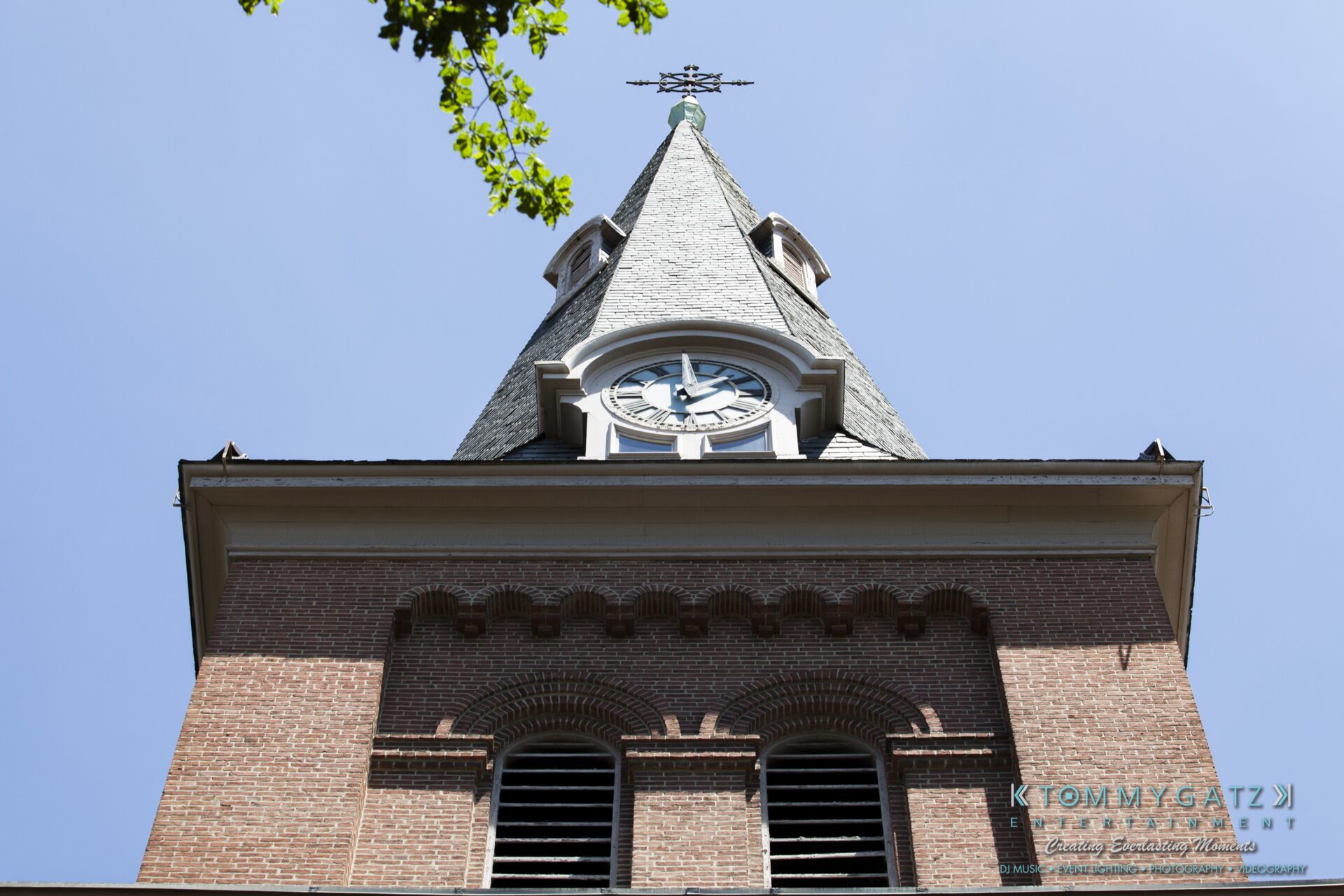 Jamie-and-Mark-Low-Res-Watermark-21 - TOMMY GATZ ENTERTAINMENT A brick church steeple with a clock face under a clear blue sky, framed by a tree branch on the left.