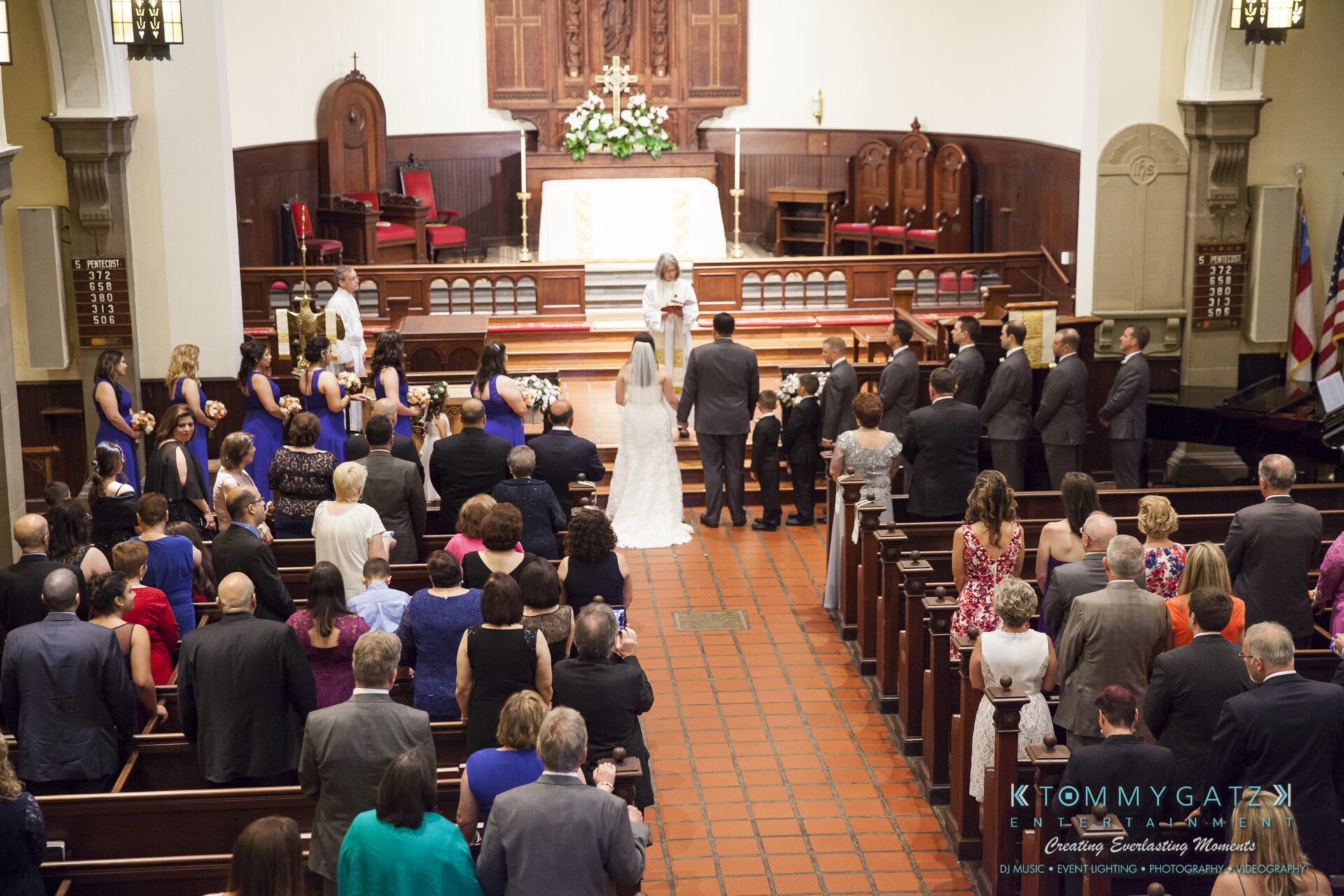 Jamie-and-Mark-Low-Res-Watermark-218 - TOMMY GATZ ENTERTAINMENT A wedding ceremony taking place in a church. The couple stands at the altar with the officiant, surrounded by guests seated in pews.