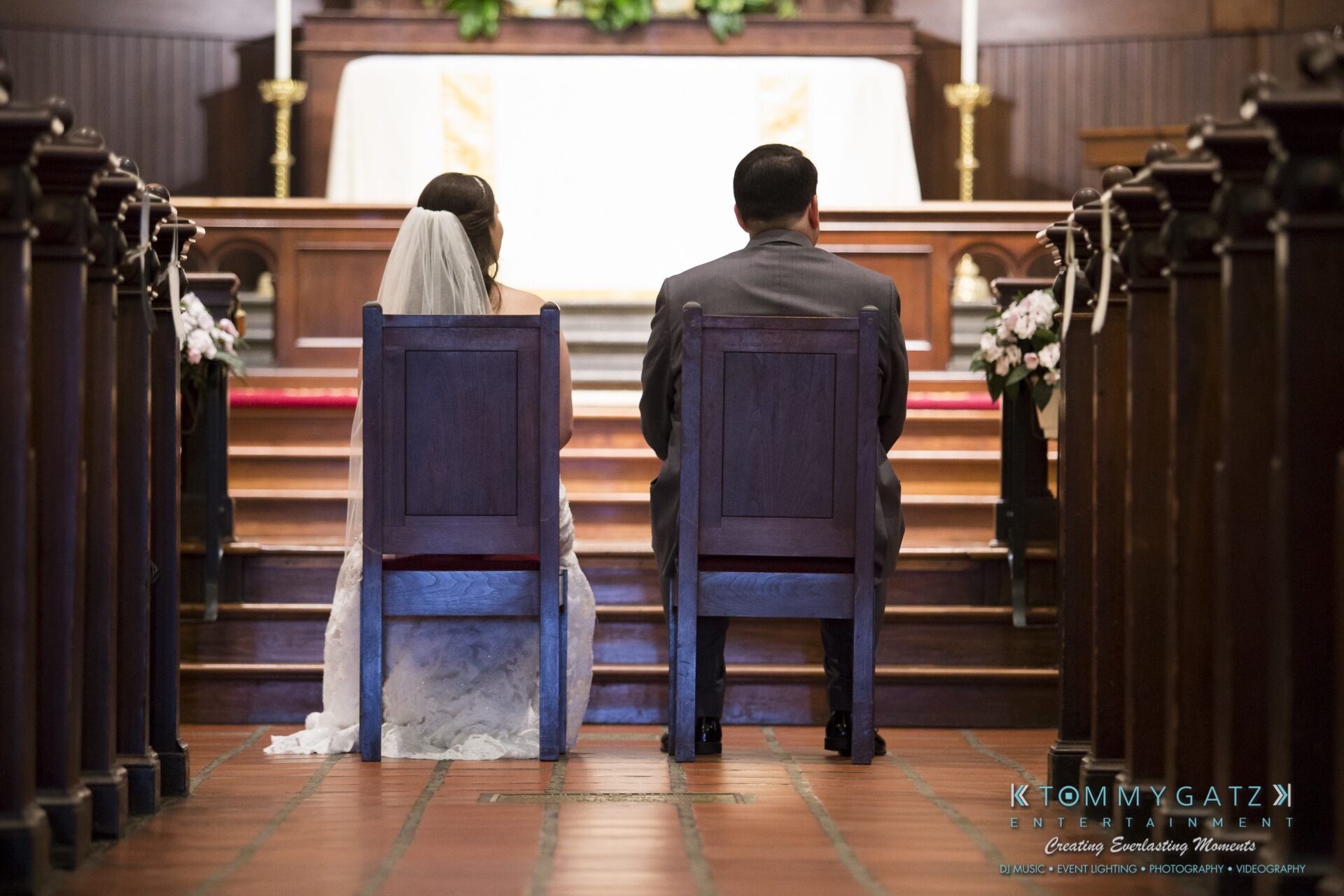 Jamie-and-Mark-Low-Res-Watermark-237 - TOMMY GATZ ENTERTAINMENT A bride and groom sit side by side in a church facing the altar, with floral arrangements visible on either side.