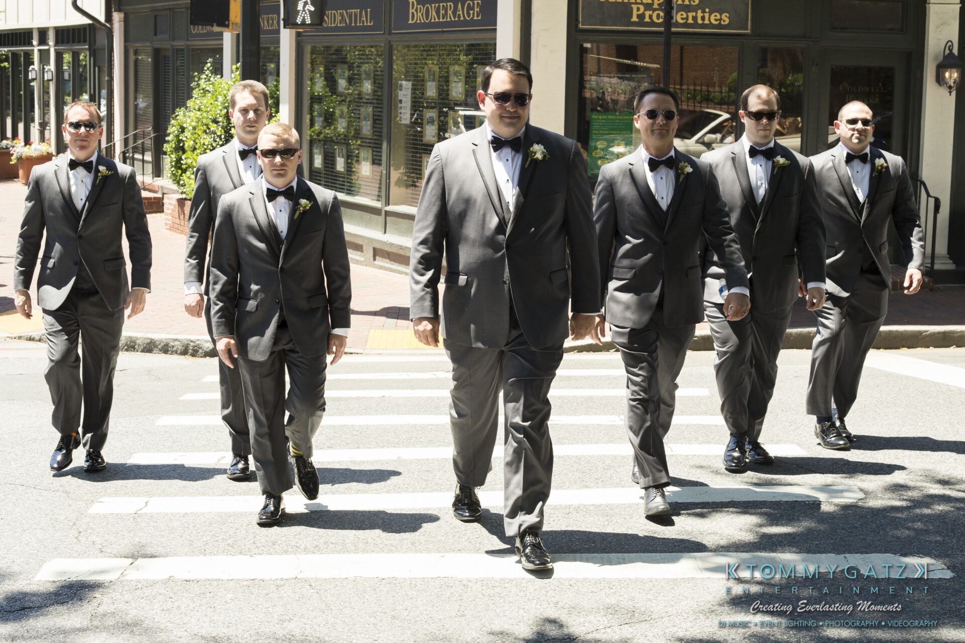 Jamie-and-Mark-Low-Res-Watermark-24 - TOMMY GATZ ENTERTAINMENT Seven men in suits and bow ties walk confidently on a crosswalk under sunlight, with storefronts in the background.