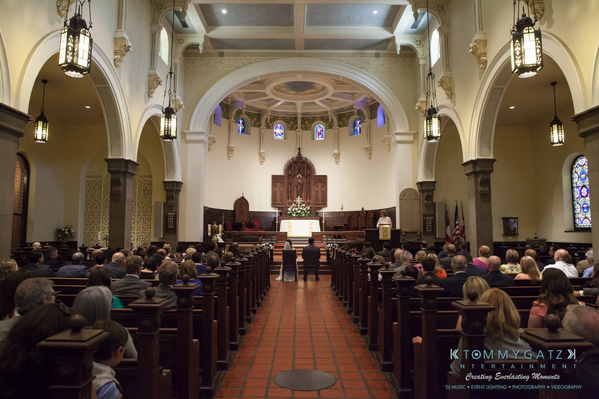 Jamie-and-Mark-Low-Res-Watermark-240 - TOMMY GATZ ENTERTAINMENT A church interior filled with people attending a formal event. The aisle is centered, leading to an altar adorned with flowers. Stained glass windows and arched ceilings are visible.