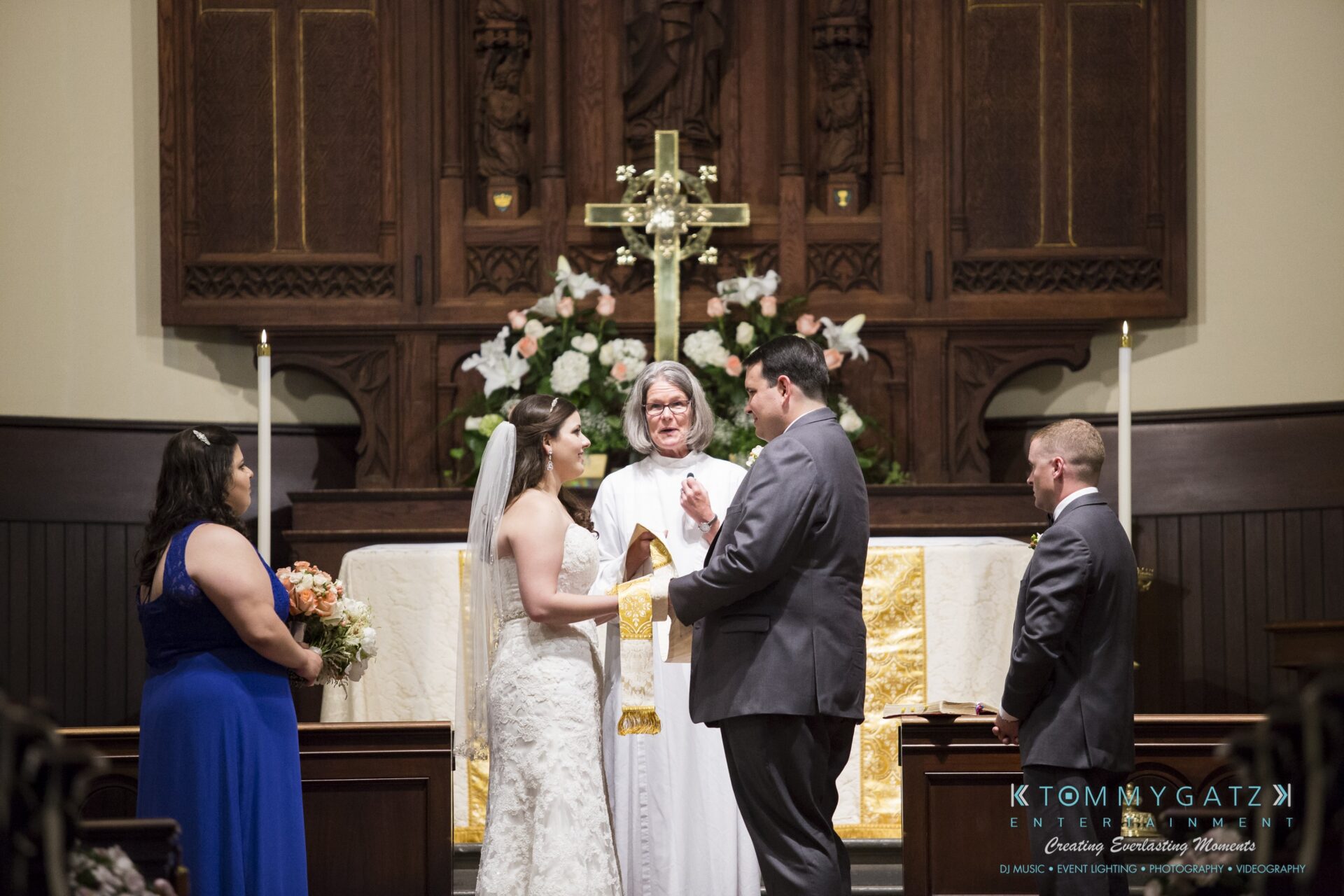 Jamie-and-Mark-Low-Res-Watermark-260 - TOMMY GATZ ENTERTAINMENT A couple exchanges vows at the altar during a wedding ceremony in a church, with a priest and bridal party members present.