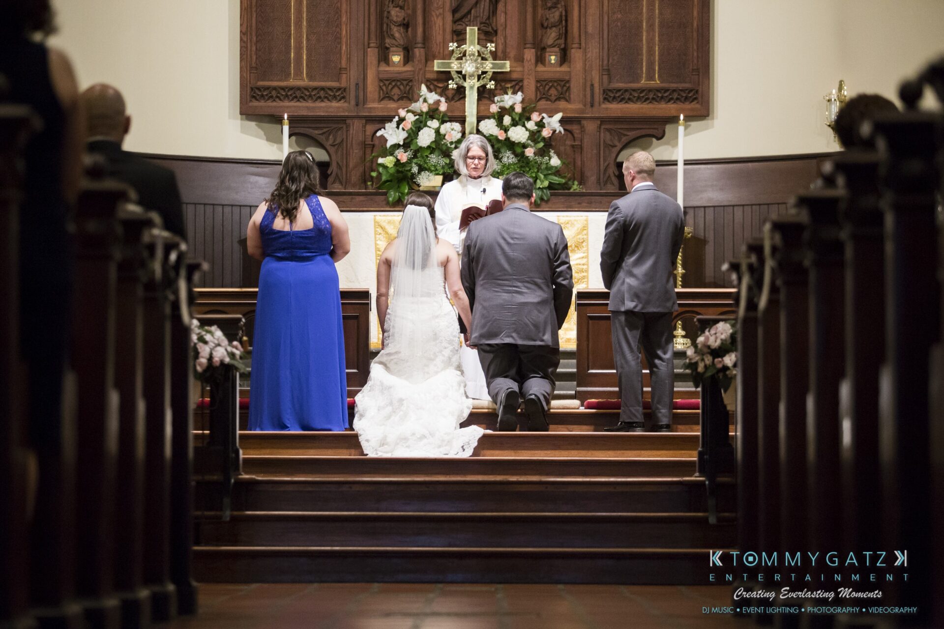 Jamie-and-Mark-Low-Res-Watermark-266 - TOMMY GATZ ENTERTAINMENT A wedding ceremony at an altar with a priest, bride, groom, and two attendants. The bride and groom kneel while the priest conducts the service.