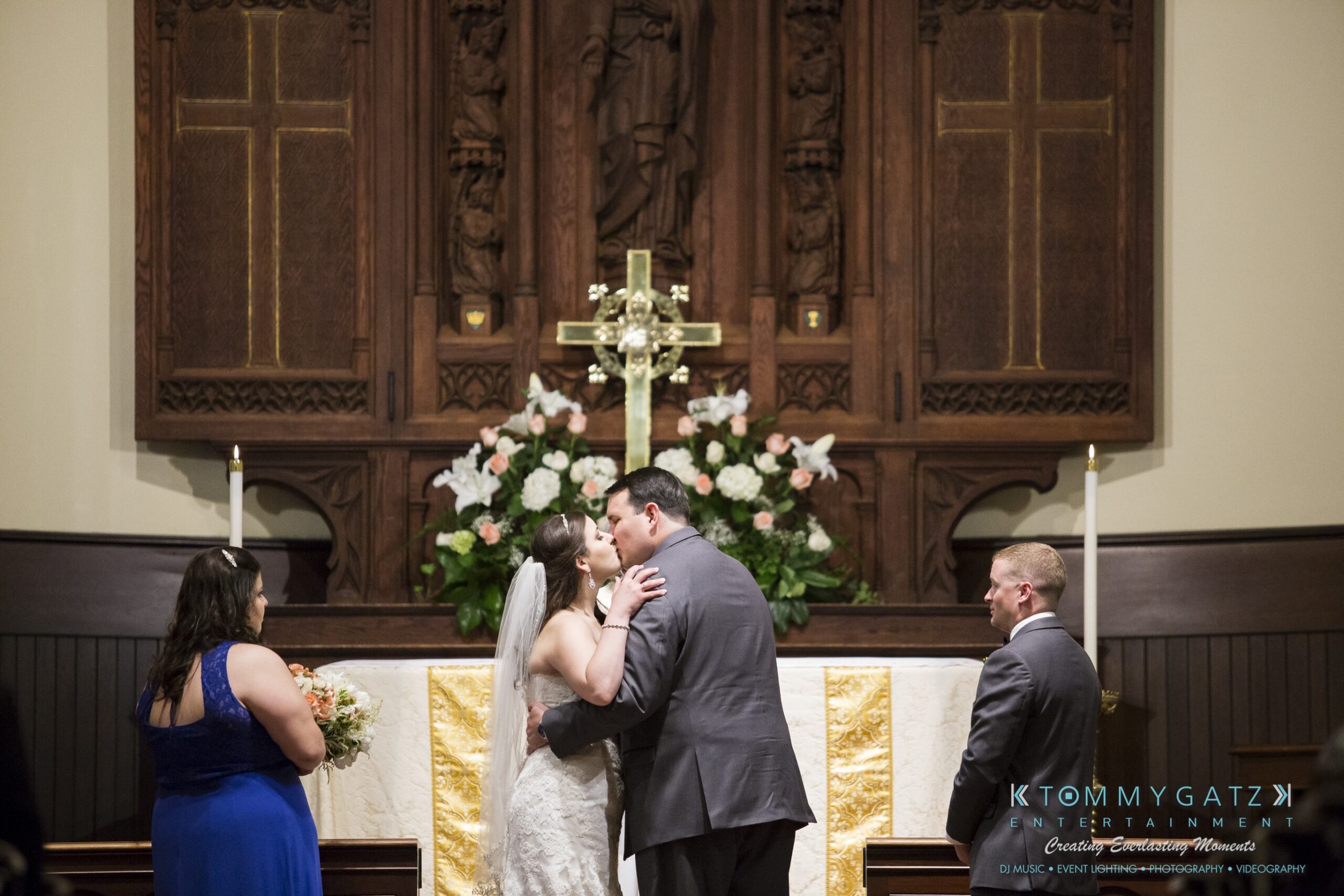 Jamie-and-Mark-Low-Res-Watermark-272 - TOMMY GATZ ENTERTAINMENT A bride and groom kiss at the altar during a wedding ceremony. Bridesmaids and groomsmen stand nearby.