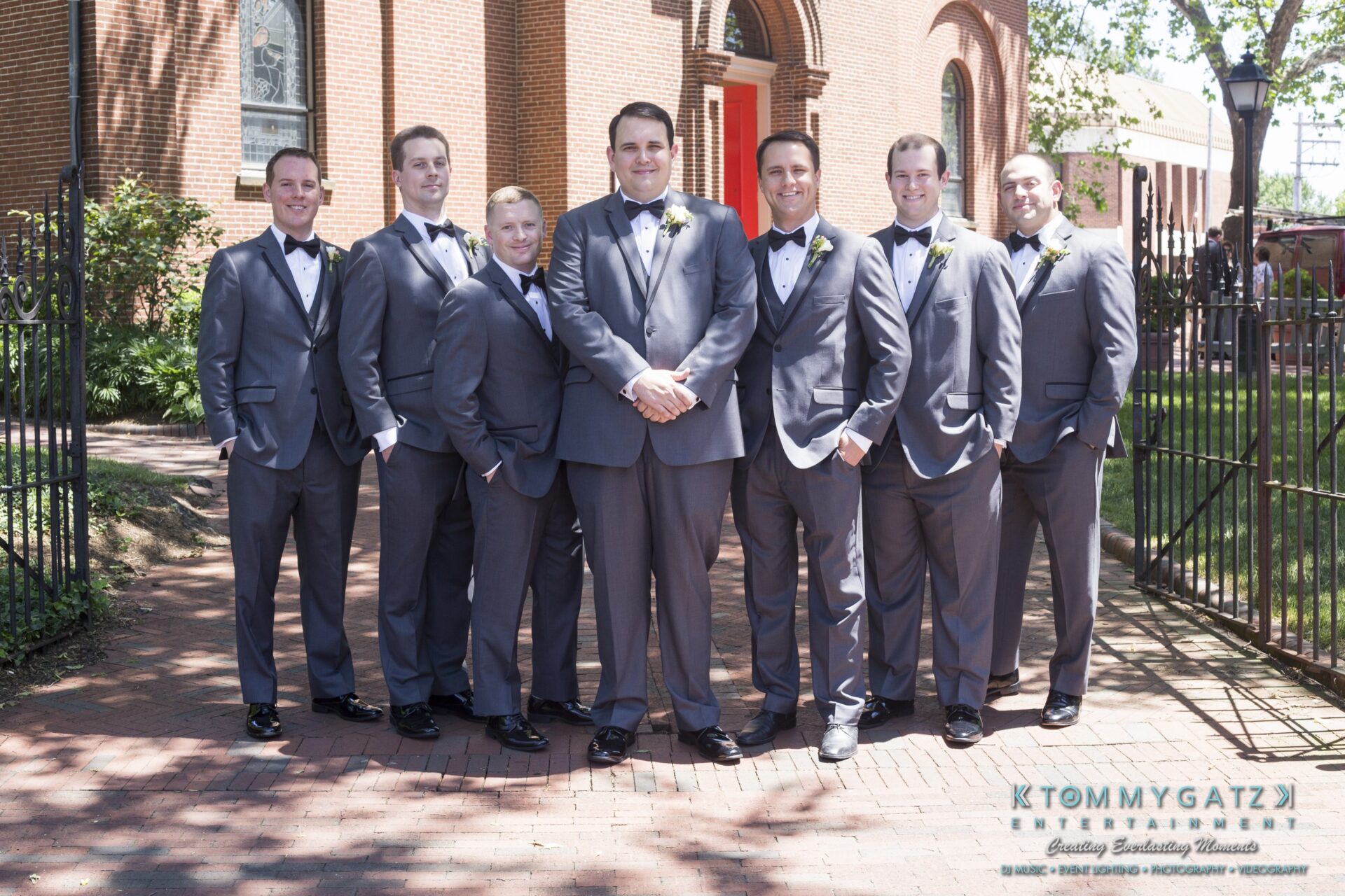 Jamie-and-Mark-Low-Res-Watermark-29 - TOMMY GATZ ENTERTAINMENT A group of seven men in gray suits and bow ties stand outdoors on a brick path, in front of a building.