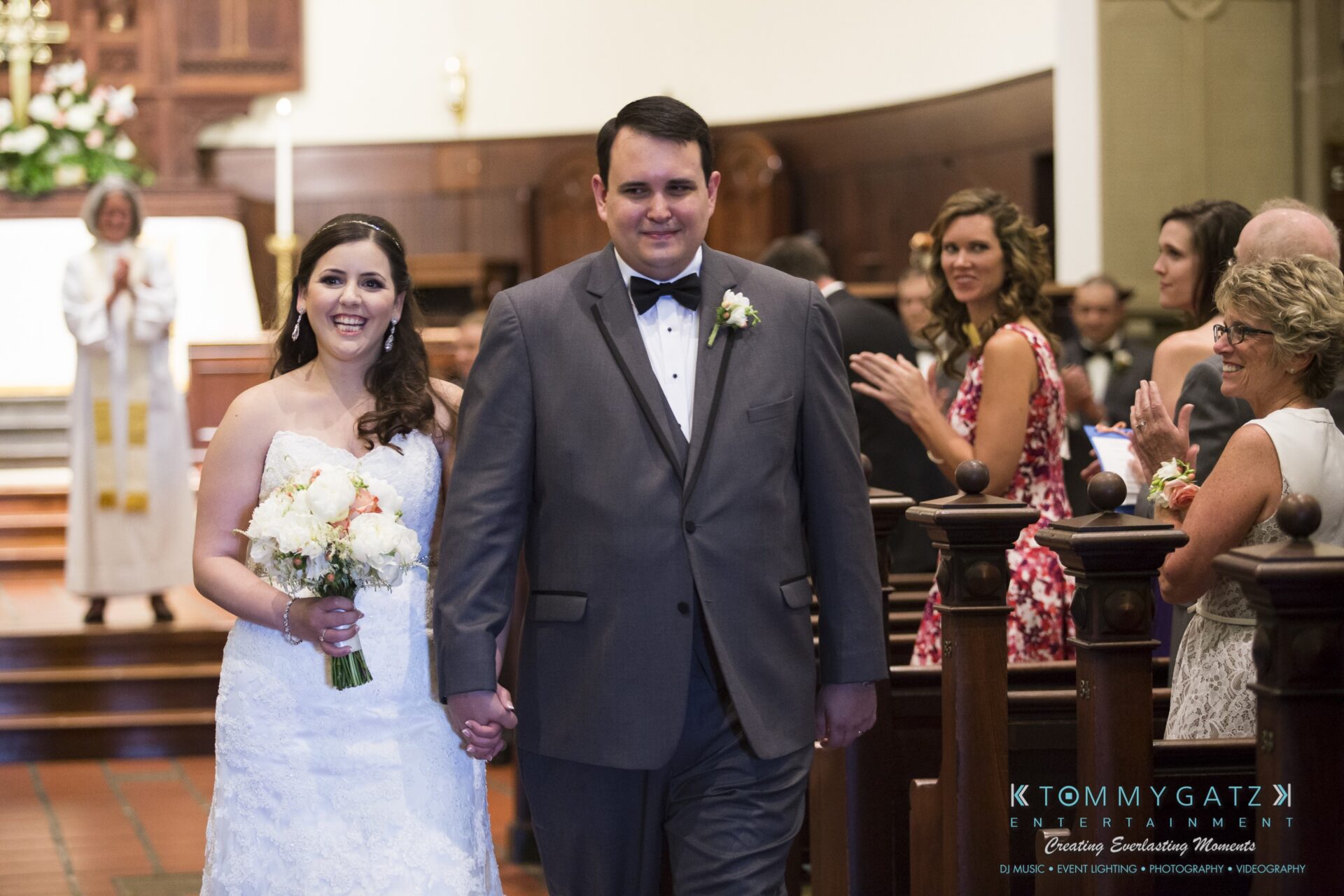 Jamie-and-Mark-Low-Res-Watermark-292 - TOMMY GATZ ENTERTAINMENT A bride and groom smile while walking down the aisle of a church as guests applaud.