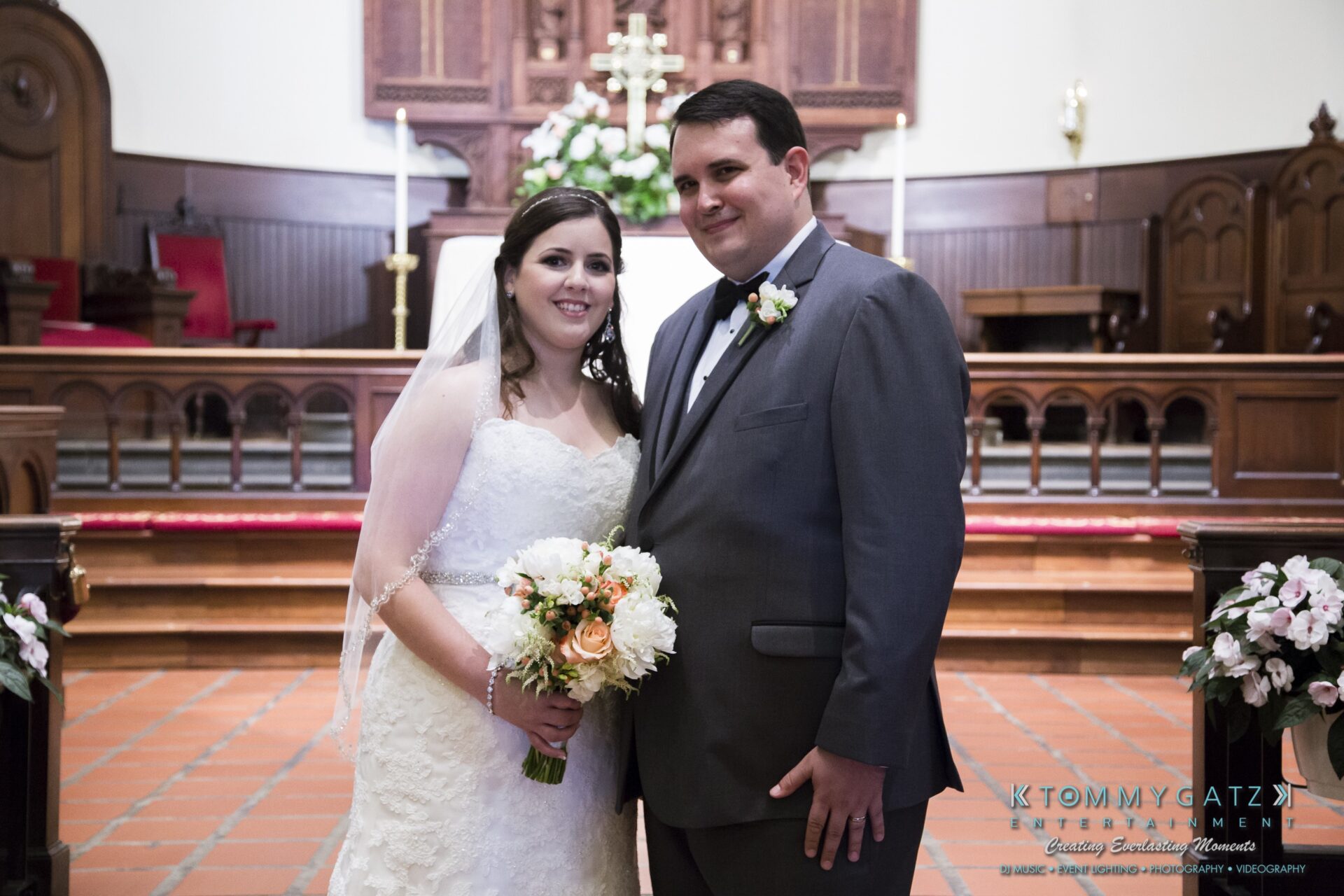 Jamie-and-Mark-Low-Res-Watermark-318 - TOMMY GATZ ENTERTAINMENT A bride and groom stand together inside a church, dressed in wedding attire, with floral decorations in the background.