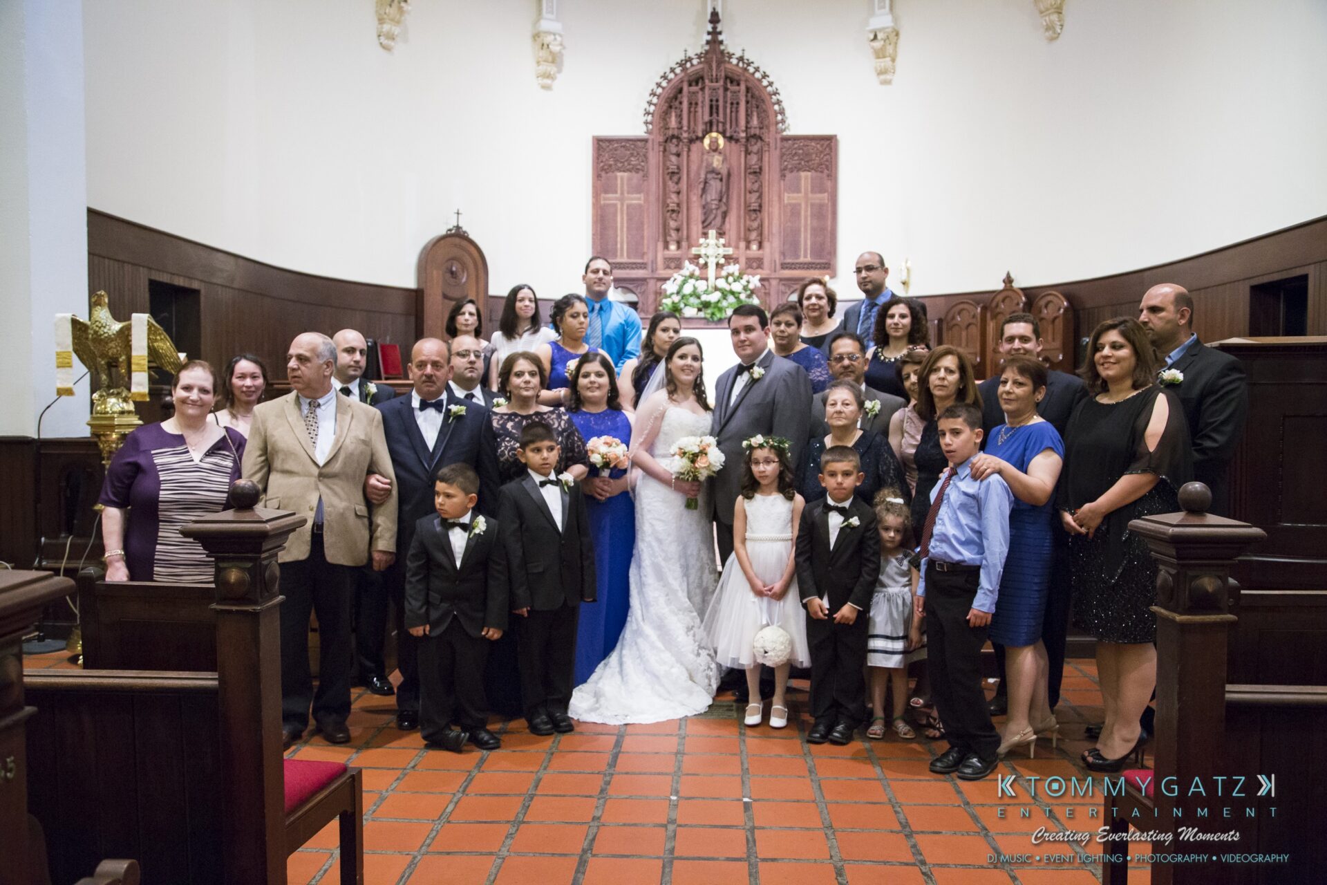 Jamie-and-Mark-Low-Res-Watermark-322 - TOMMY GATZ ENTERTAINMENT A large wedding party poses inside a church. The bride and groom stand centered, surrounded by family and friends, with an altar in the background.